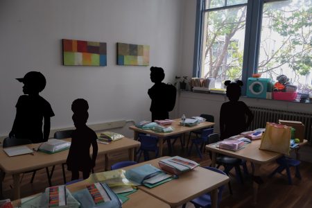A classroom with four desks, school supplies neatly organized in bins, large window, colorful artwork on the wall, and silhouettes of four children standing and sitting.