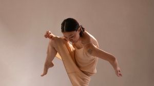 A person in a beige outfit performs a dance movement, raising one knee and extending their arms, against a neutral background.