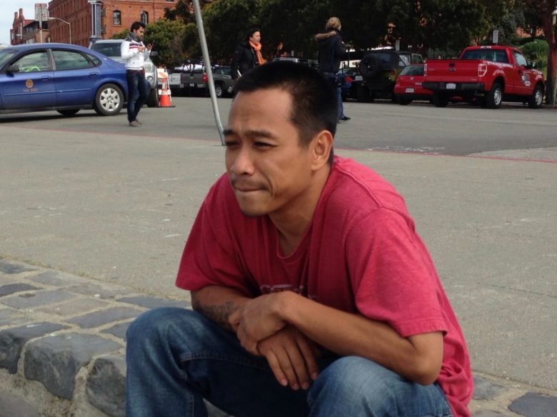 A man in a red shirt and jeans sits on a curb with his hands clasped, looking thoughtful. Cars and people are visible in the background.