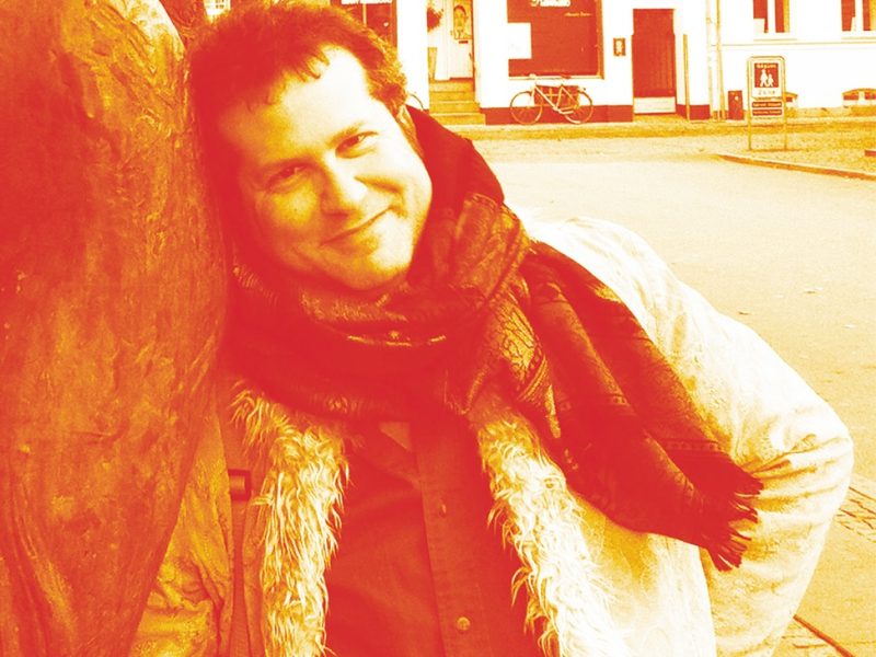 A person wearing a scarf and coat leans against a large statue outdoors, smiling at the camera on a city street.