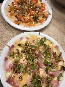 Two plates of sashimi are shown; one features sliced fish topped with herbs and sauce, the other has cherry tomatoes, herbs, and a light dressing. Both are placed on a wooden table.
