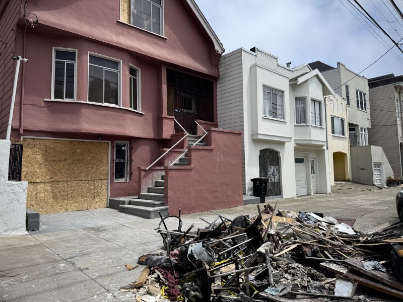 A pile of debris and broken furniture lies on the sidewalk in front of a row of houses, one of which has boarded-up windows.
