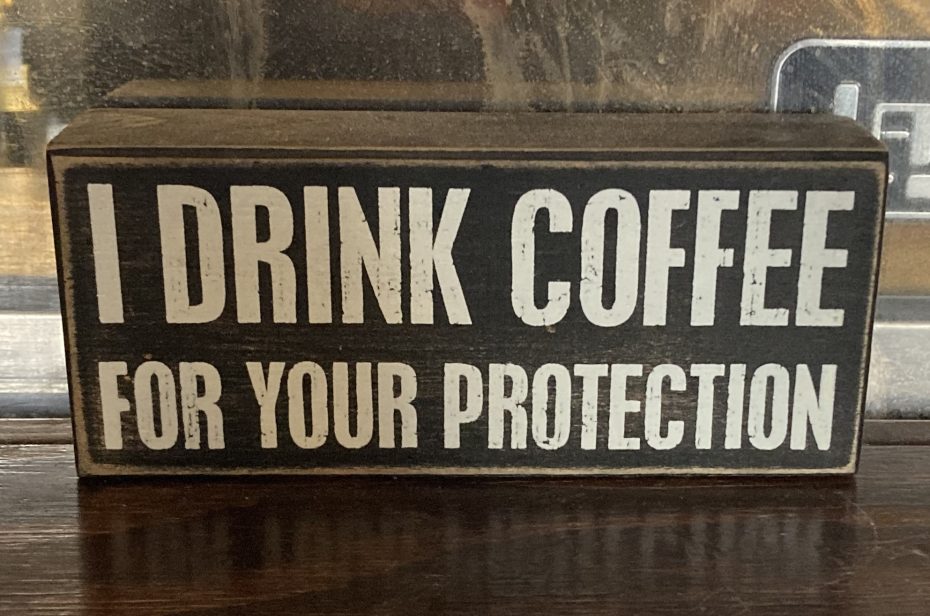 A rectangular wooden sign with bold white text reads, "I DRINK COFFEE FOR YOUR PROTECTION.
