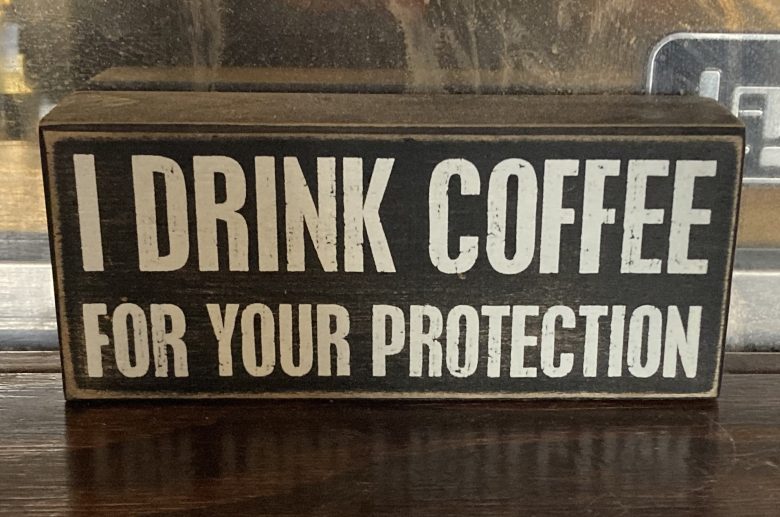 A wooden sign with bold white text reading, "I DRINK COFFEE FOR YOUR PROTECTION," displayed on a dark surface.