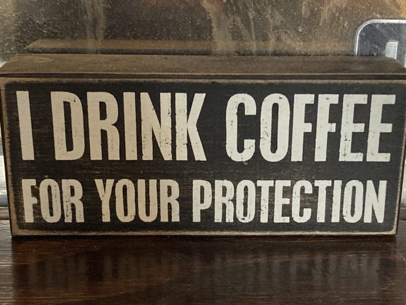 A wooden sign with bold white text reading, "I DRINK COFFEE FOR YOUR PROTECTION," displayed on a dark surface.