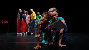 A group of people in colorful clothing perform on stage, with two individuals kneeling at the front and a digital clock reading "5:00" in the background.