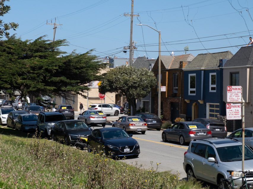 Cars with yellow signs along a residential street lined with houses; "No Parking" signs are posted nearby under a clear blue sky.