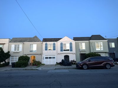 A row of attached houses with garages at street level, a red SUV parked on the right, and a motorcycle covered in the center driveway at dusk.