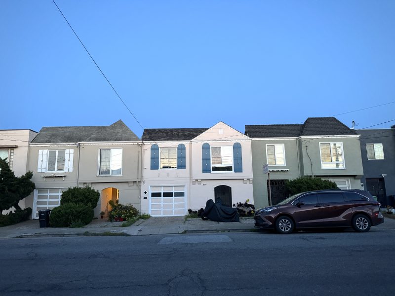 A row of attached houses with garages at street level, a red SUV parked on the right, and a motorcycle covered in the center driveway at dusk.