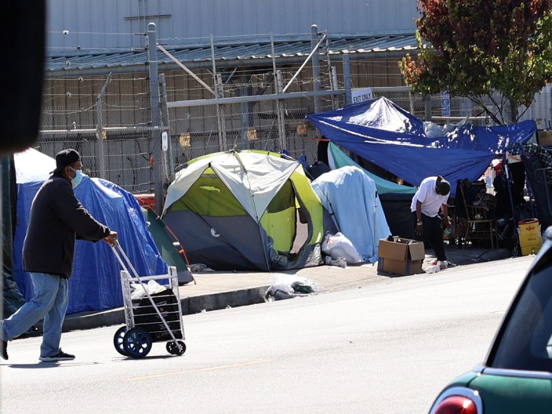 A man pushes a cart past tents and shelters set up on a sidewalk, with belongings and makeshift structures visible along a fenced area.