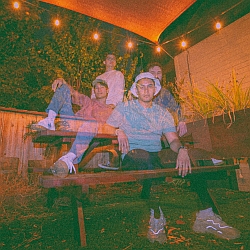 Four people sit and stand around a wooden picnic table outdoors at night under string lights, with a double exposure effect creating a ghostly appearance.