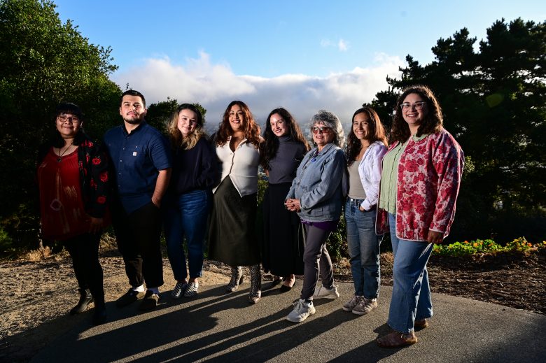 Eight people stand side by side outdoors on a paved path with trees and a cloudy sky in the background.