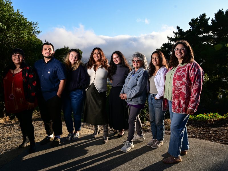 Eight people stand side by side outdoors on a paved path with trees and a cloudy sky in the background.