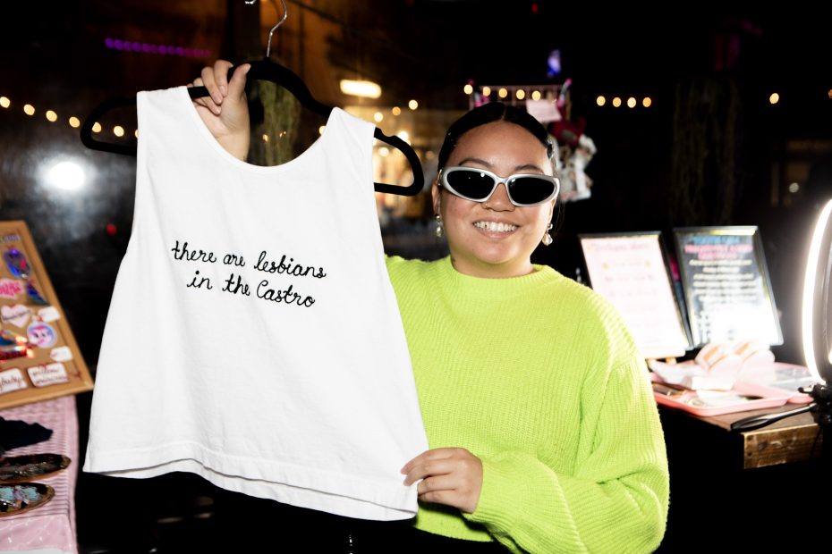 A person wearing sunglasses and a neon green sweater holds up a white tank top that reads, "there are lesbians in the Castro.