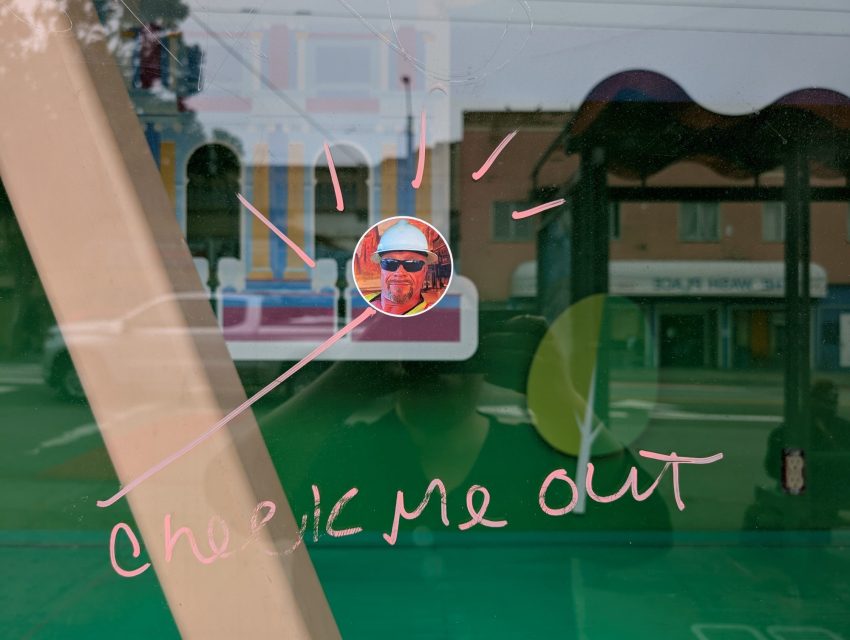 Photo of a window with a round sticker showing a man in sunglasses and a hard hat, pink marker text reads "check me out" with lines drawn to the sticker.