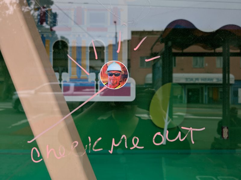 A sticker of a man wearing sunglasses and a hat is on a window, with "check me out" written in pink marker and lines radiating from the sticker.