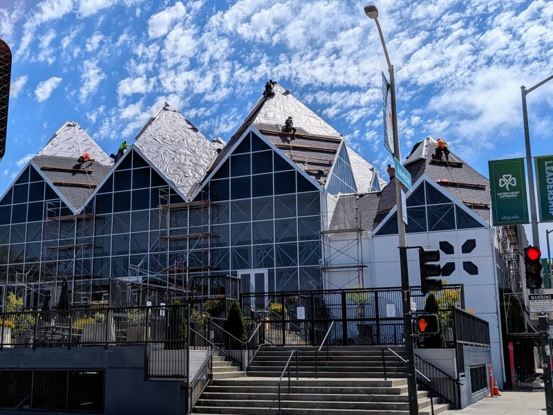 Workers install new roofing materials on the angular, glass-fronted roof of a modern building under a partly cloudy sky. Scaffolding is set up around the structure.