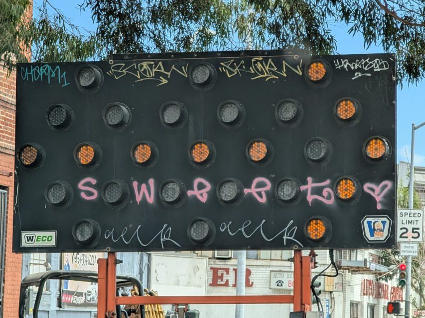 Electronic road sign with some amber lights lit up, covered in graffiti tags and "sweet" spray-painted in pink, on a city street.