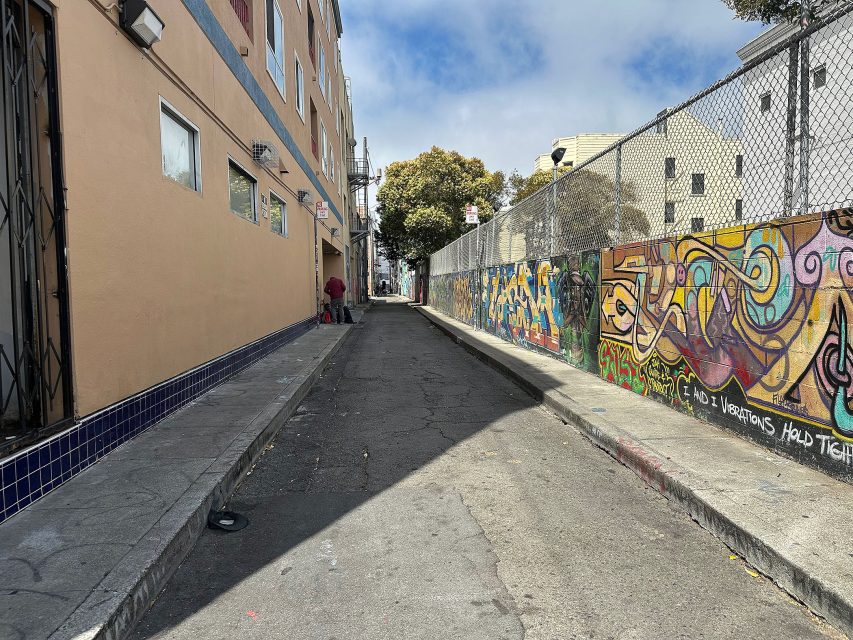 A narrow urban alleyway with colorful graffiti on a fence to the right and a beige building to the left, under a partly cloudy sky.