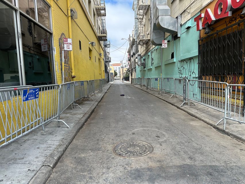 A narrow urban alleyway with metal barricades on both sides, yellow and green buildings, graffiti, and empty of people.