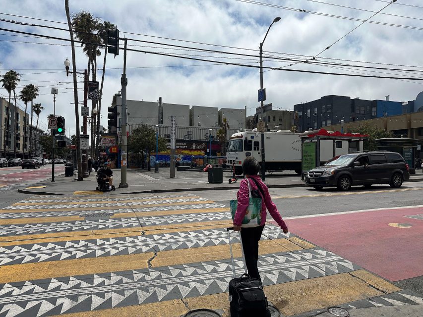 A person with a suitcase waits to cross a city street at a crosswalk with geometric patterns; traffic and buildings are in the background.