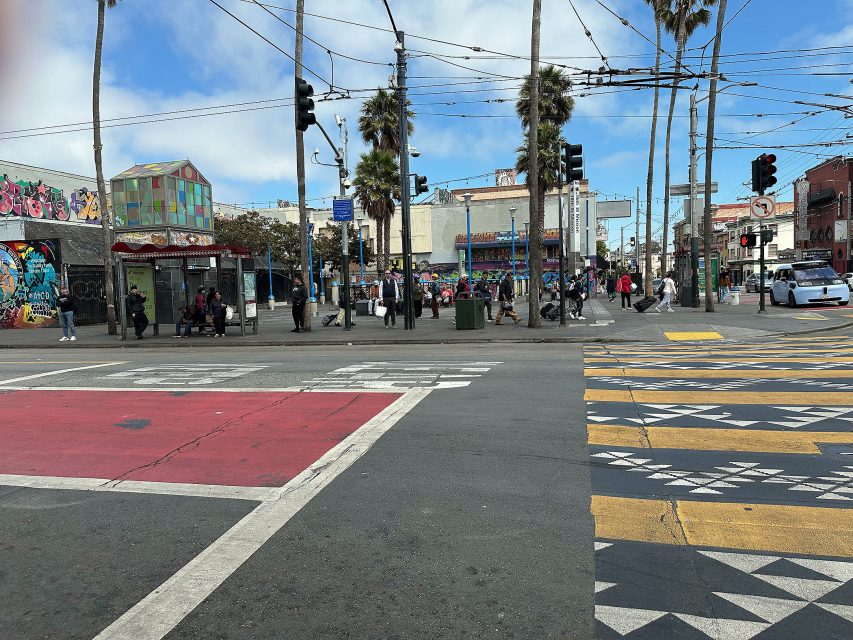 Urban intersection with crosswalks, people waiting, palm trees, street art, traffic signals, and overhead wires under a partly cloudy sky.