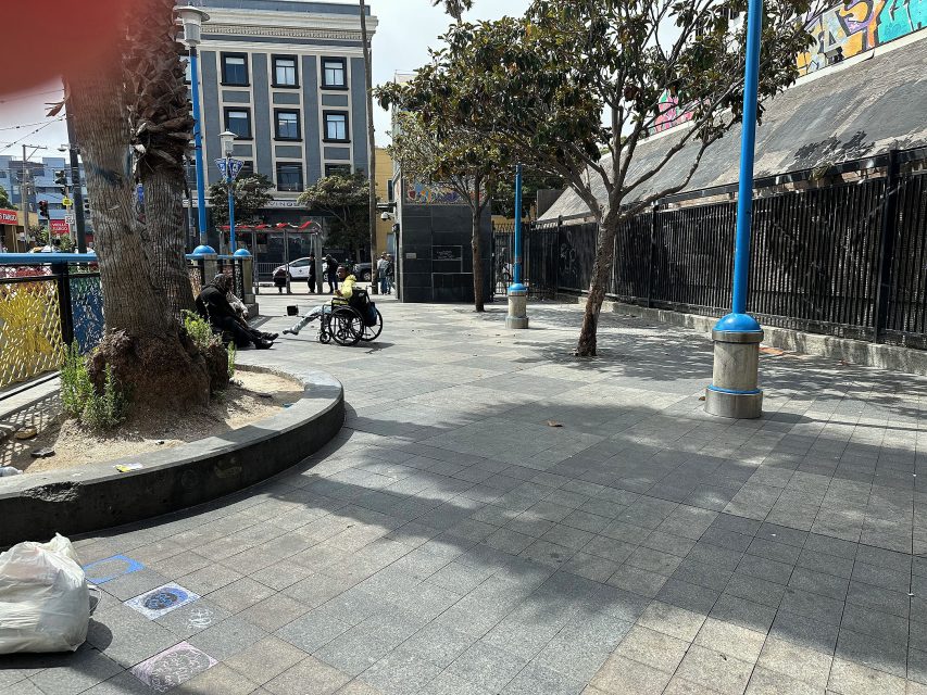 A person in a wheelchair and another person sit by trees in an urban plaza with blue lamp posts, buildings, and a fenced area in the background.