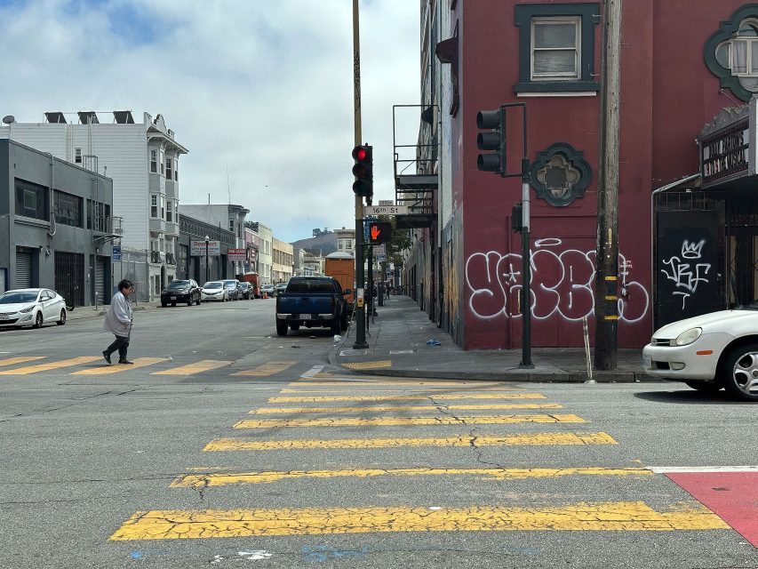 A person crosses a street at a crosswalk on an urban corner with traffic signals, parked cars, and graffiti on the building wall.