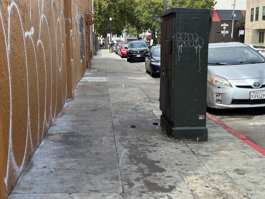 A city sidewalk with graffiti on the wall, parked cars along the street, and a utility box near the curb. Trees and buildings are visible in the background.