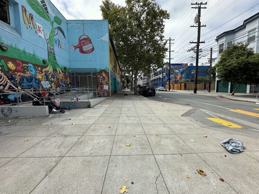A city sidewalk with scattered debris, mural-covered buildings, a tree, parked cars, and overhead power lines under a cloudy sky.