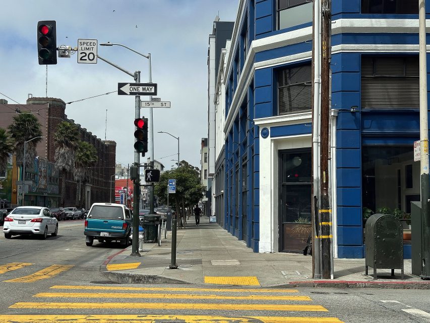 Street intersection with traffic lights showing red, a blue and white building on the corner, crosswalks, cars stopped, and clear street signs visible.