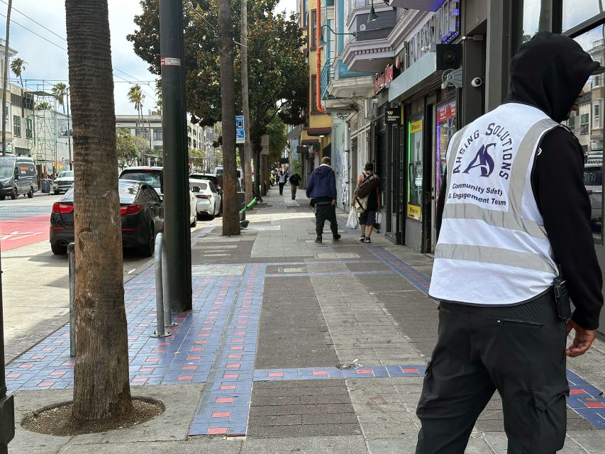 A person wearing a “Safety Solutions” vest stands on a city sidewalk, with several people walking and cars parked along the street.