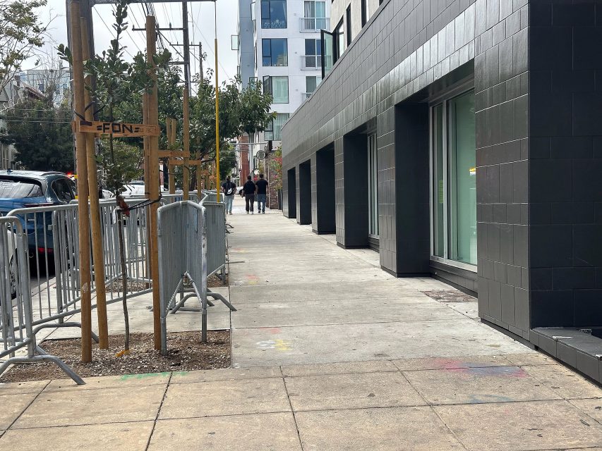 A city sidewalk with metal barriers, a small tree, and two people walking in the distance near modern buildings.