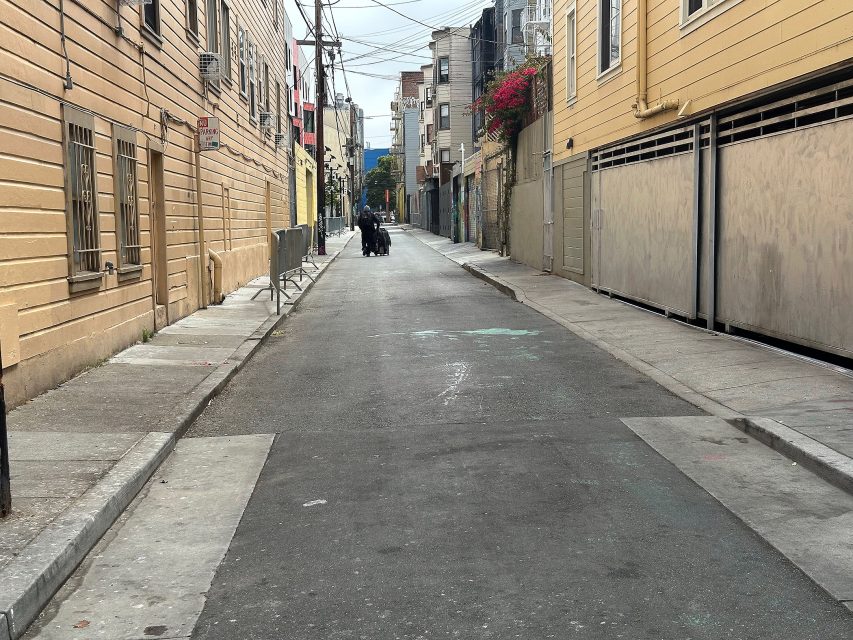 A narrow urban alley with beige buildings on both sides, some graffiti, a person walking away in the distance, and overhead utility wires crossing above.