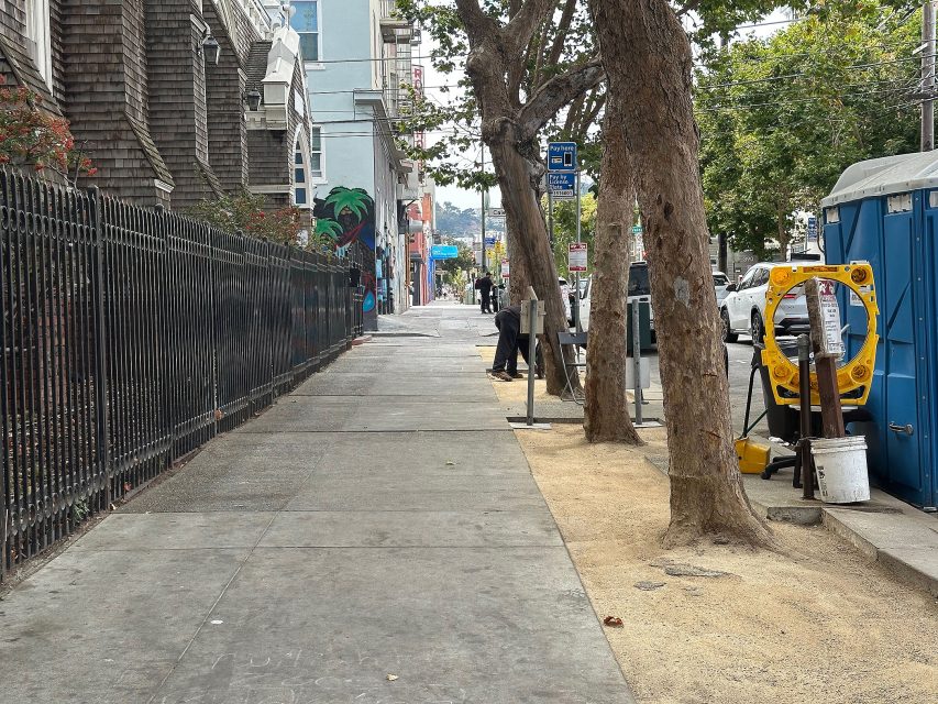A city sidewalk with trees, a fenced building on the left, a portable toilet, and construction tools on the right. A few people are visible in the background.