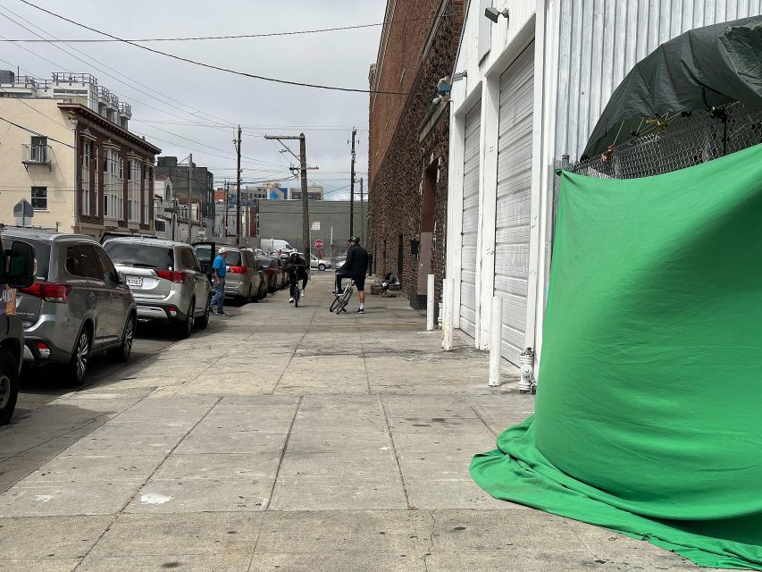 A city street with parked cars, two people walking in the distance, and a large green screen draped over a fence on the right side of the sidewalk.