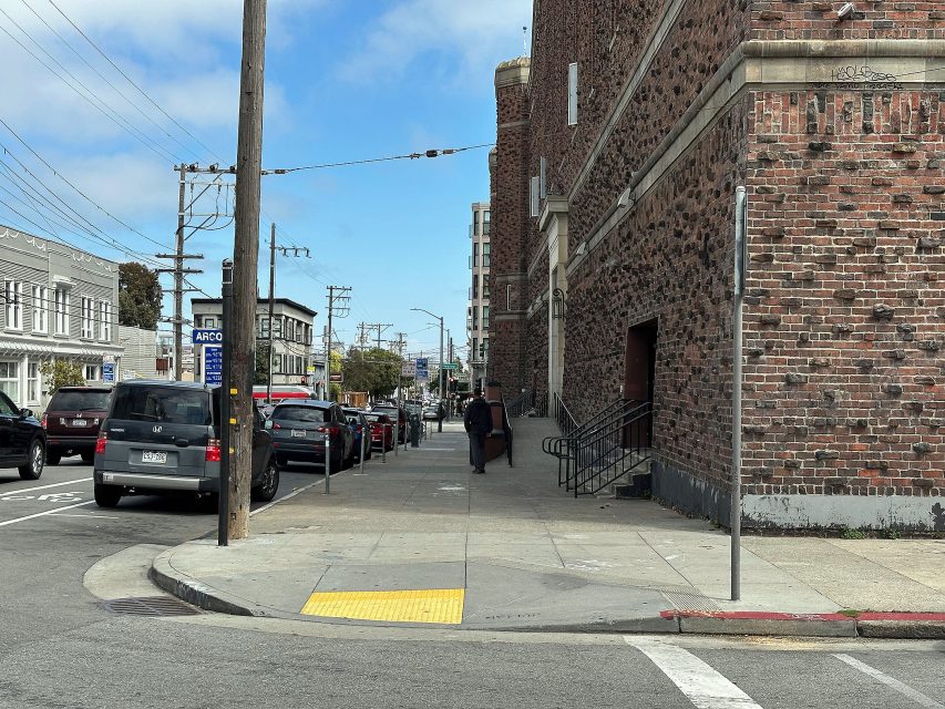 A city sidewalk with parked cars, a bus stop sign, a brick building, and two people walking away from the camera under a partly cloudy sky.