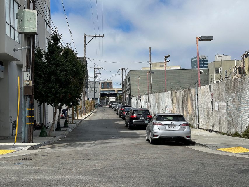 A narrow urban street with parked cars along one side, bordered by buildings and a tall concrete wall, under a partly cloudy sky.