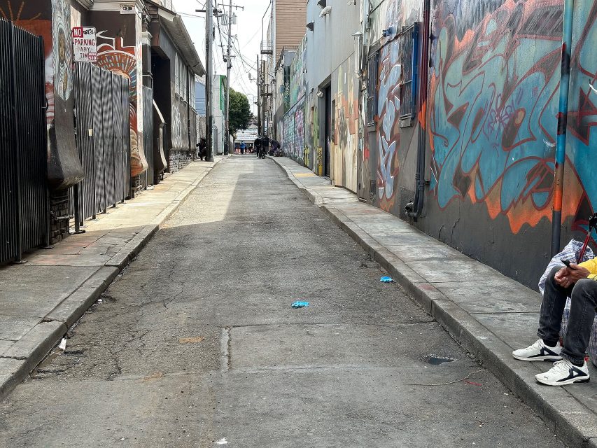 A narrow urban alleyway with graffiti-covered walls, scattered debris on the ground, and a person sitting on the right edge of the image.