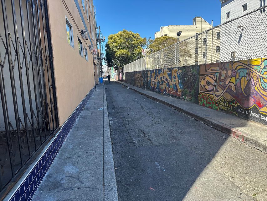 Narrow urban alleyway bordered by a building and a fence with colorful graffiti, under a clear blue sky.