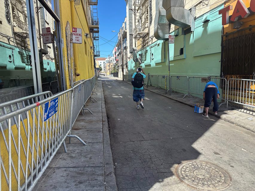 Two people walk down a narrow alley with metal barricades, a yellow building on the left, and a person crouched near a paint can on the right.