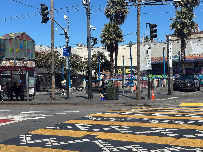 A city street corner with a colorful crosswalk, palm trees, people waiting at a bus stop, and graffiti-covered buildings in the background under a clear blue sky.