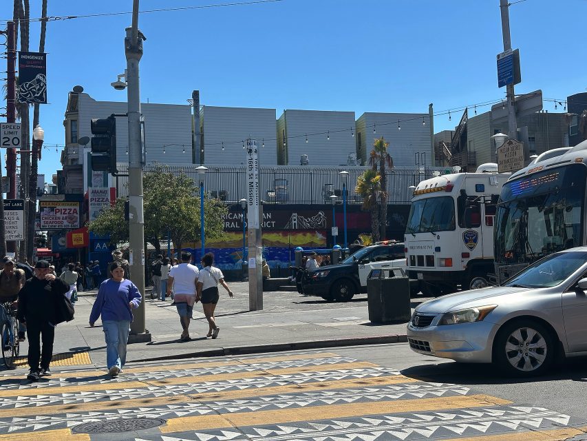 A city street scene with pedestrians crossing, parked police vehicles, and various storefronts under a clear blue sky.