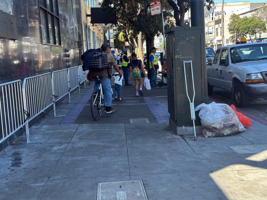A man rides a bicycle on a city sidewalk past a group of people in safety vests. A crutch leans against a utility box near a bag of trash on the ground.