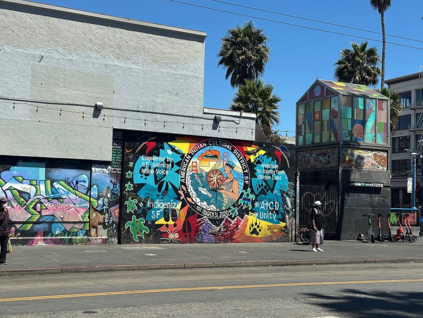 A colorful mural on a building wall features cultural symbols and the text "Indigenize SF" and "#LCD Unity"; nearby, a geometric glass structure and parked scooters line the street.