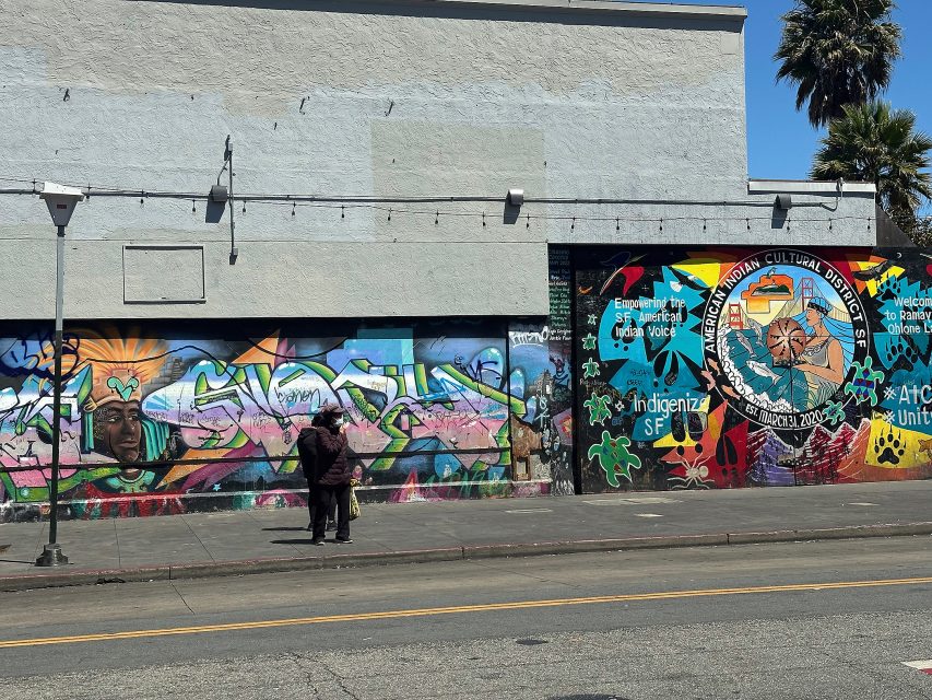 Two people stand on a sidewalk in front of a building covered in colorful murals, including Indigenous themes and phrases like "Indigenize SF" and "American Indian Cultural District.