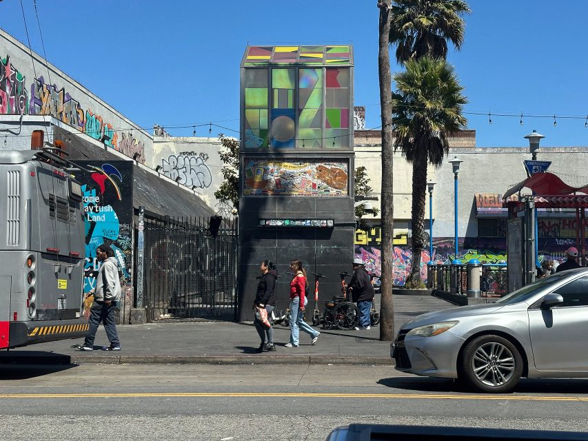 People cross a street in an urban area with graffiti-covered walls, a colorful cube sculpture, a bus, and cars visible on a sunny day.