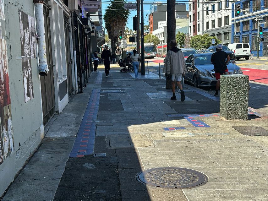 City sidewalk with pedestrians, parked cars, a manhole cover, and buildings; street art and posters visible on the left wall.