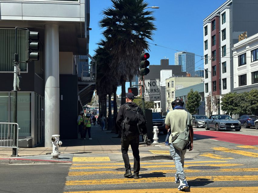 Two people cross a city street at a crosswalk under a red traffic light, with buildings, palm trees, and cars in the background on a sunny day.