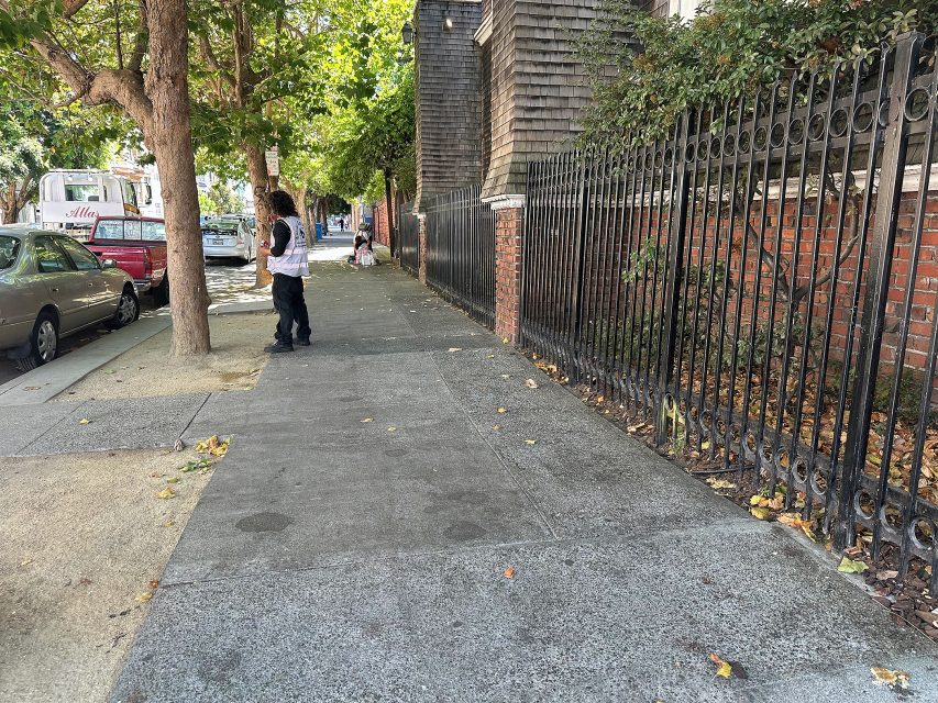 A person stands on a shaded sidewalk near a black metal fence and brick building; another person sits further down the sidewalk, with parked cars and trees lining the street.